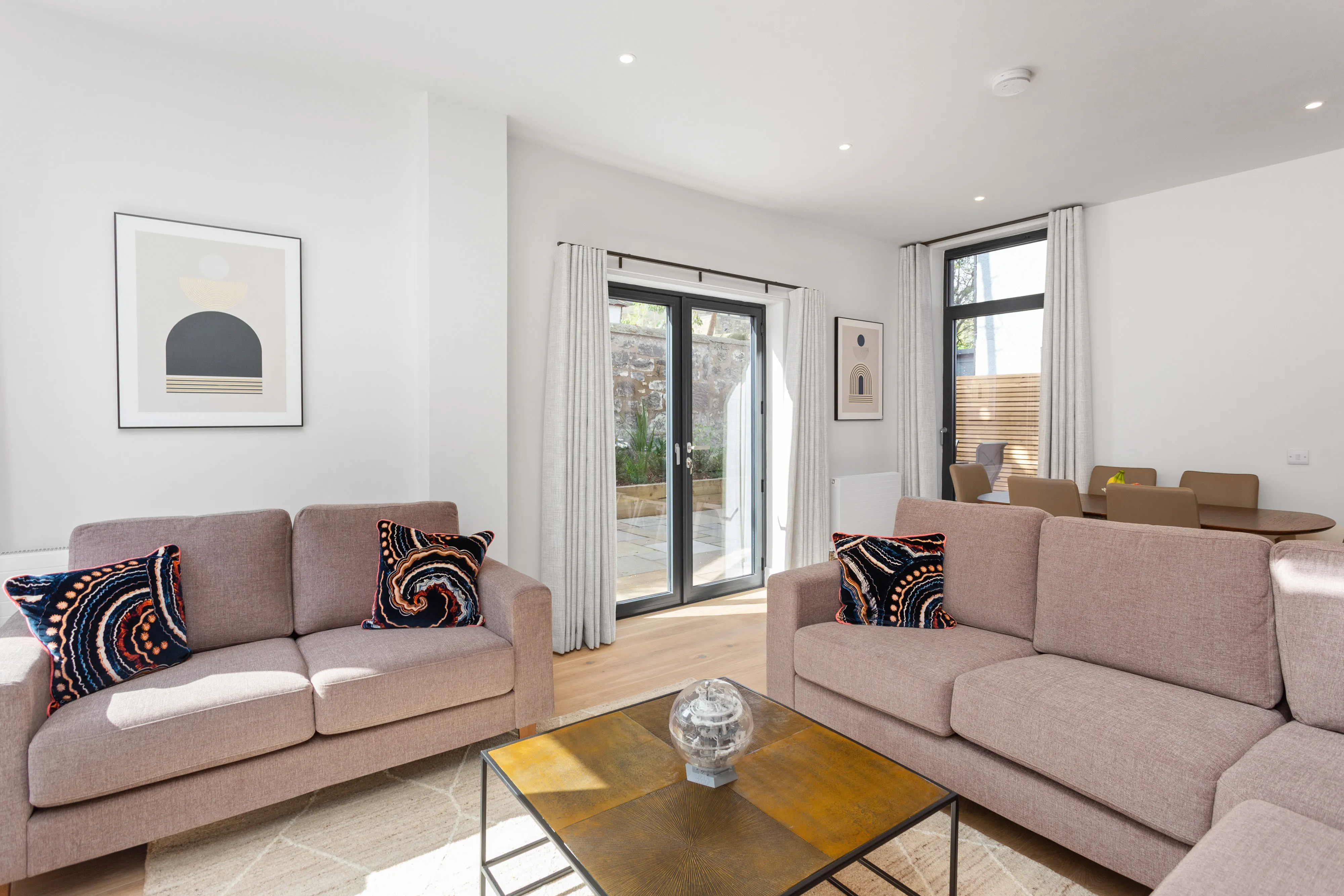 Open-plan living/dining room with two dusty-pink sofas and patterned cushions, cream curtains either side of glazed garden doors, and a walnut dining table beyond.