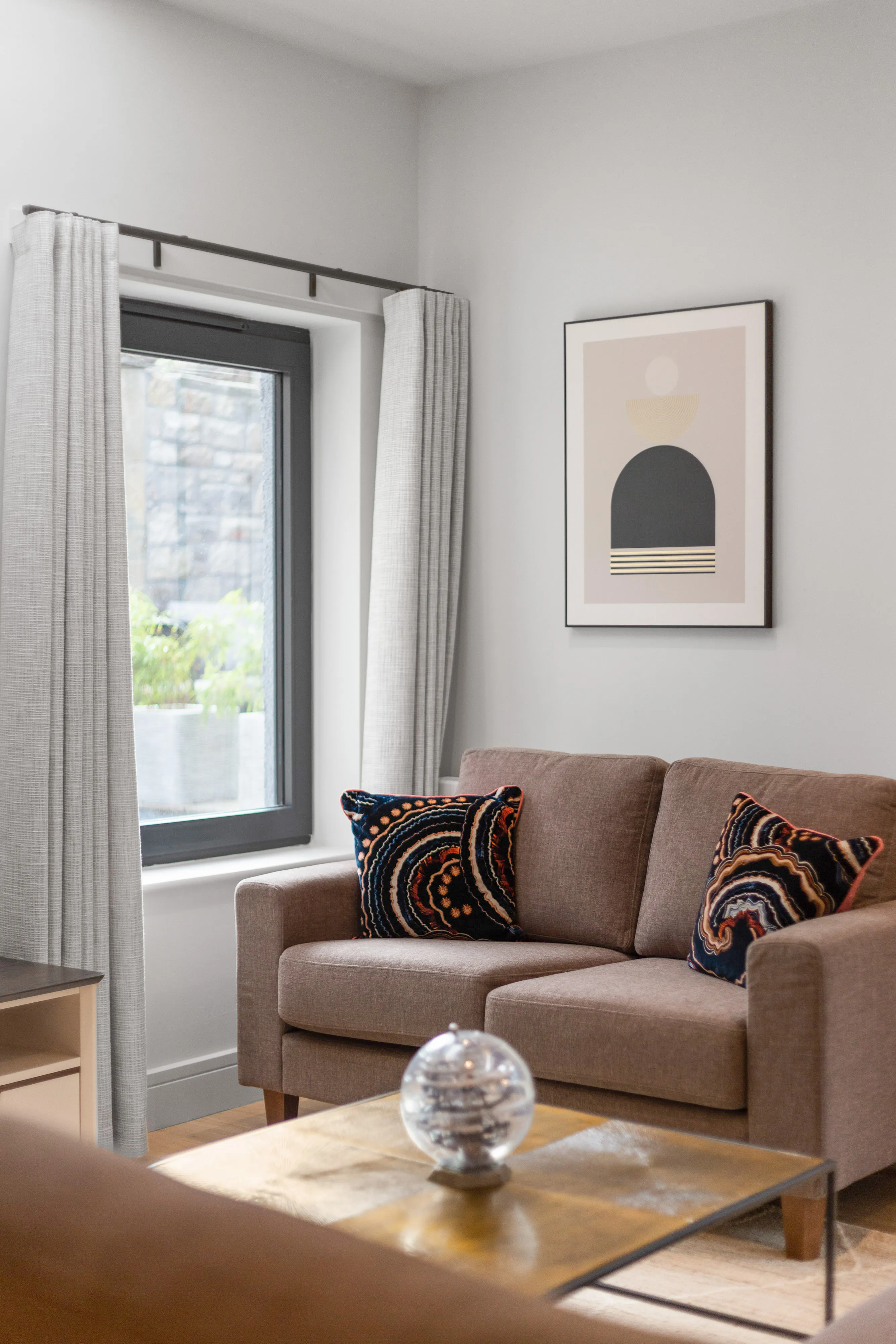 Corner of a living room with a two-seat taupe sofa and patterned cushions beside a curtained window, a single framed print on the wall above, and a glass coffee table in the foreground.