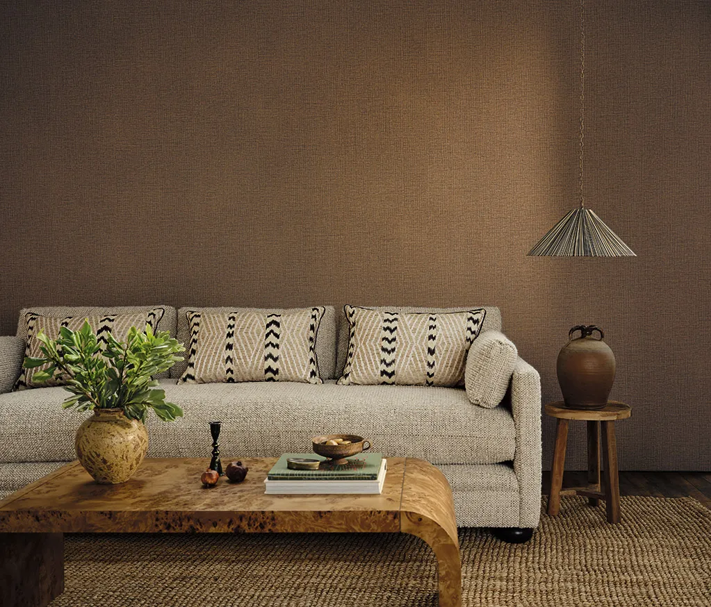 Putty-textured sofa with black and white geometric-pattern cushions against a warm brown textured wall, beside a burl-wood coffee table, a woven pendant light and a jute rug.