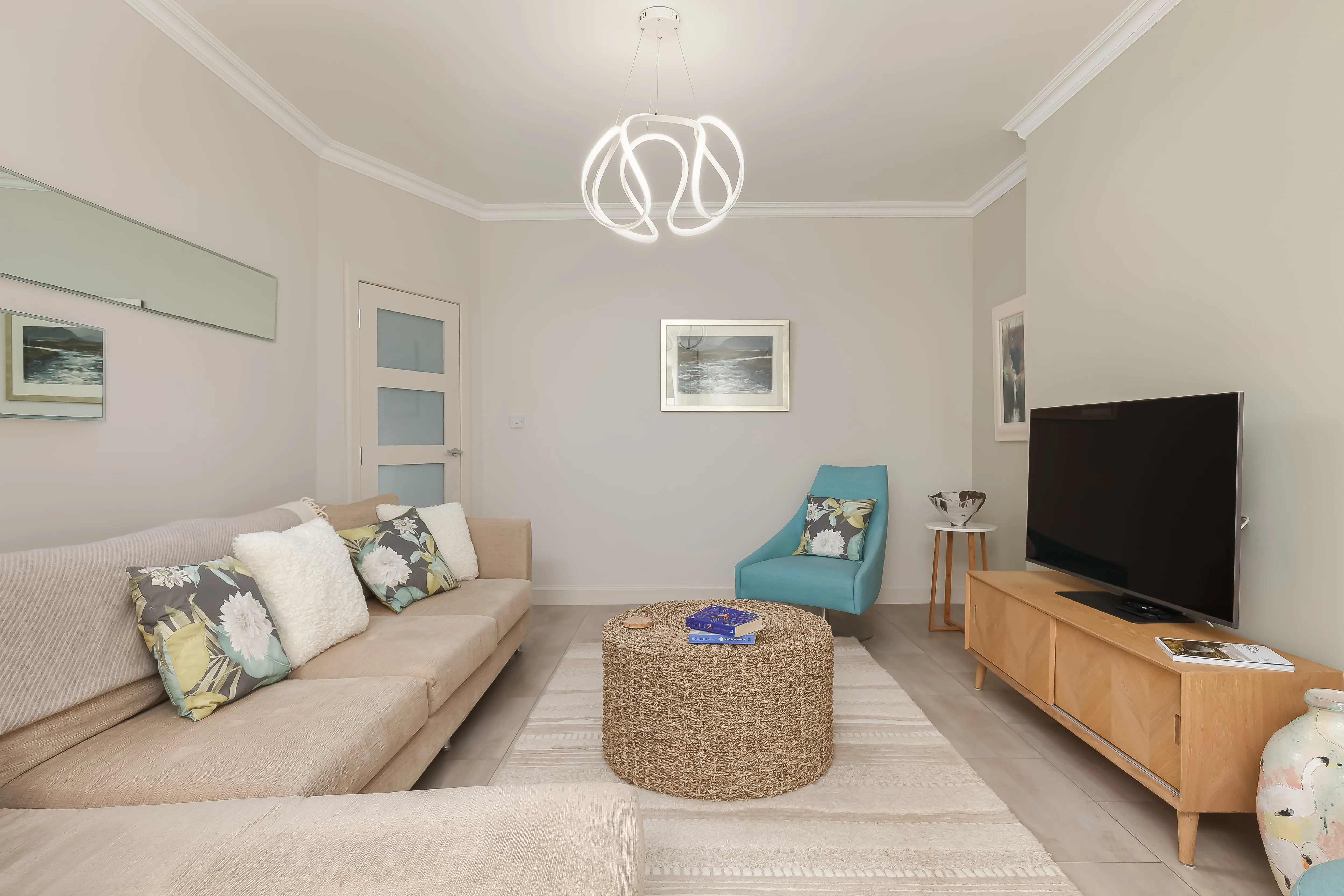 Living room with a beige corner sofa in patterned cushions, a teal accent chair, a woven-stool coffee table and a wood TV console, under a sculptural ring pendant light.