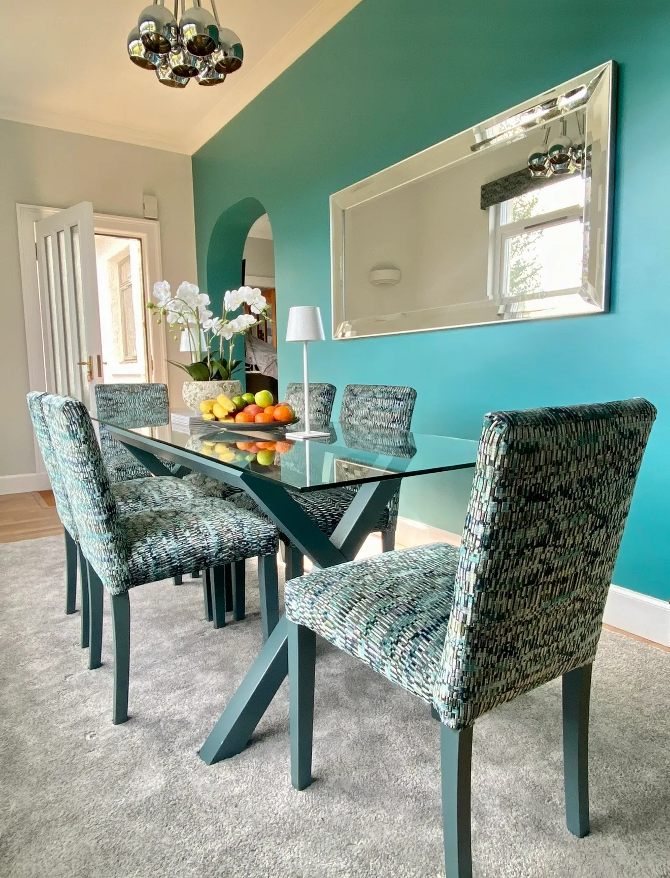 Teal dining room with a glass-topped table, six mosaic-velvet chairs, a large bevelled mirror and an arch through to the hallway.