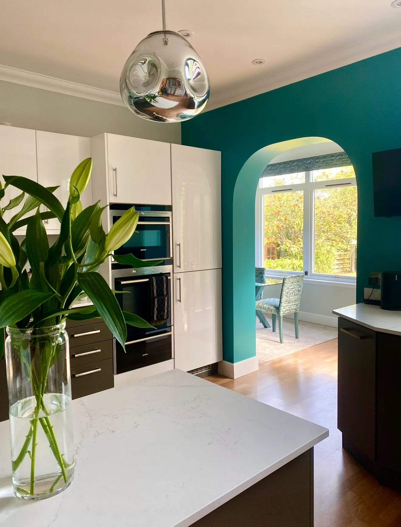 Kitchen with white gloss units, Siemens ovens, a marble-effect island and a chrome pendant, opening through a teal arch to a garden-facing dining space.