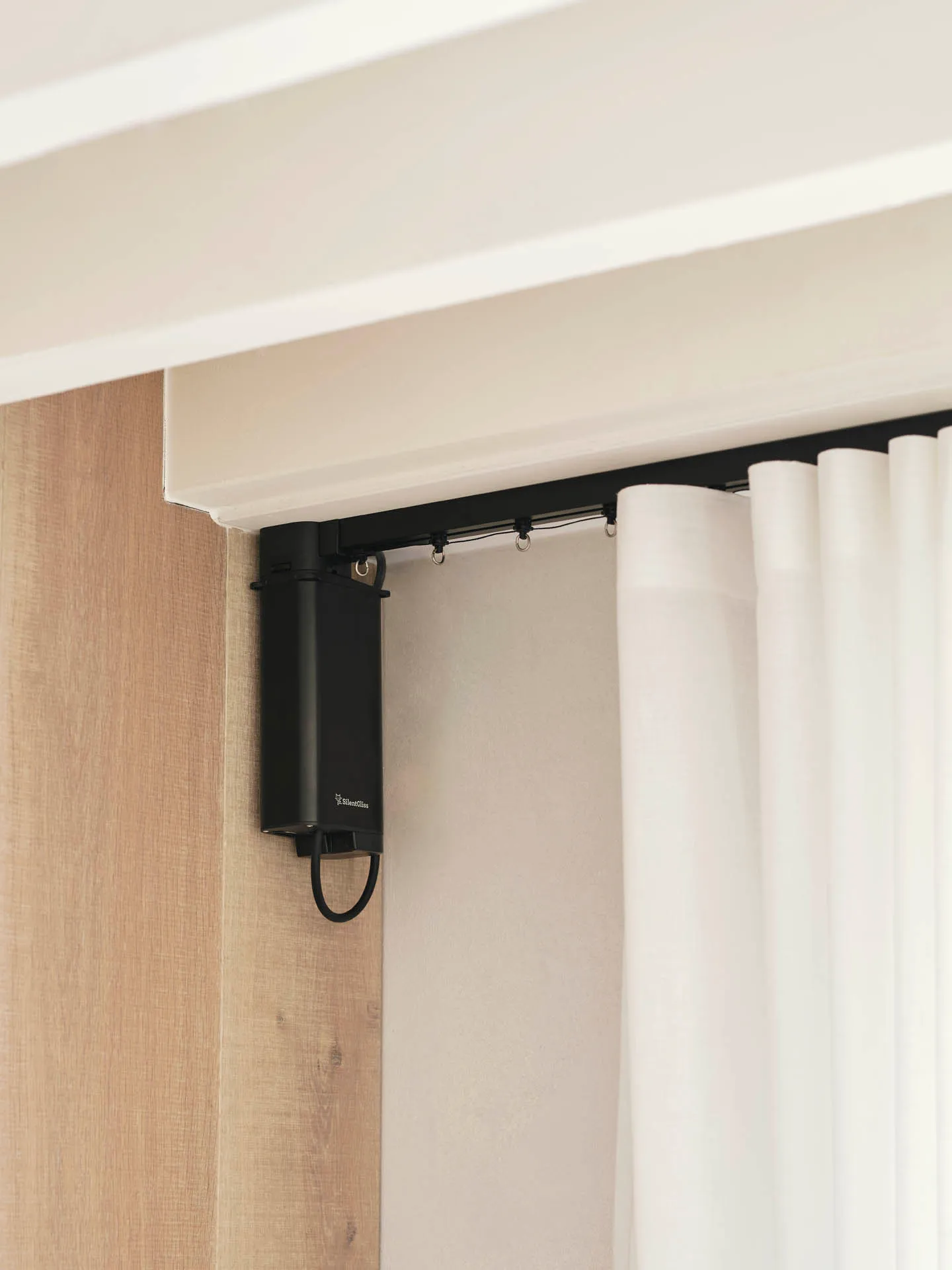 Black Silent Gliss electric curtain track with its battery motor unit, carrying a white sheer wave curtain beside pale timber panelling.