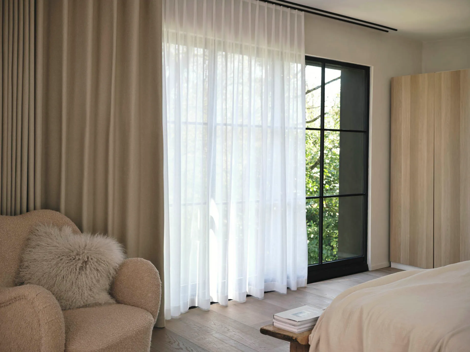 Bedroom with layered window dressing — a taupe wave over-curtain and white sheers at a black-framed window — with a bouclé armchair and a pale linen bed.