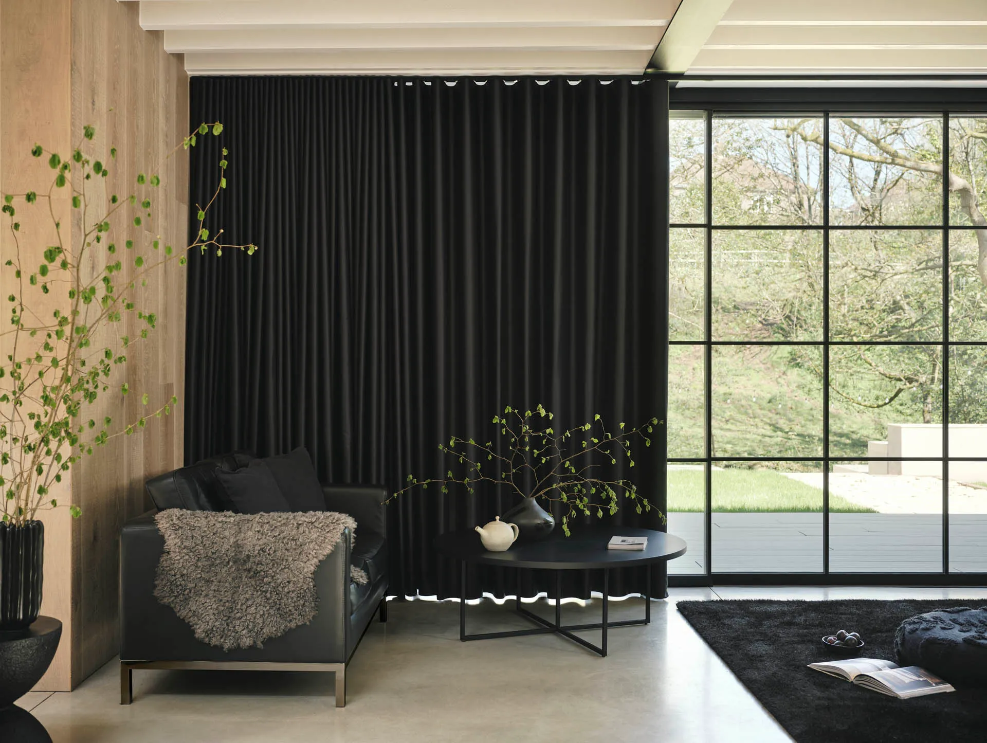 Living room with a full-height black wave curtain, a black leather sofa and a round coffee table beside floor-to-ceiling glazing to a garden.