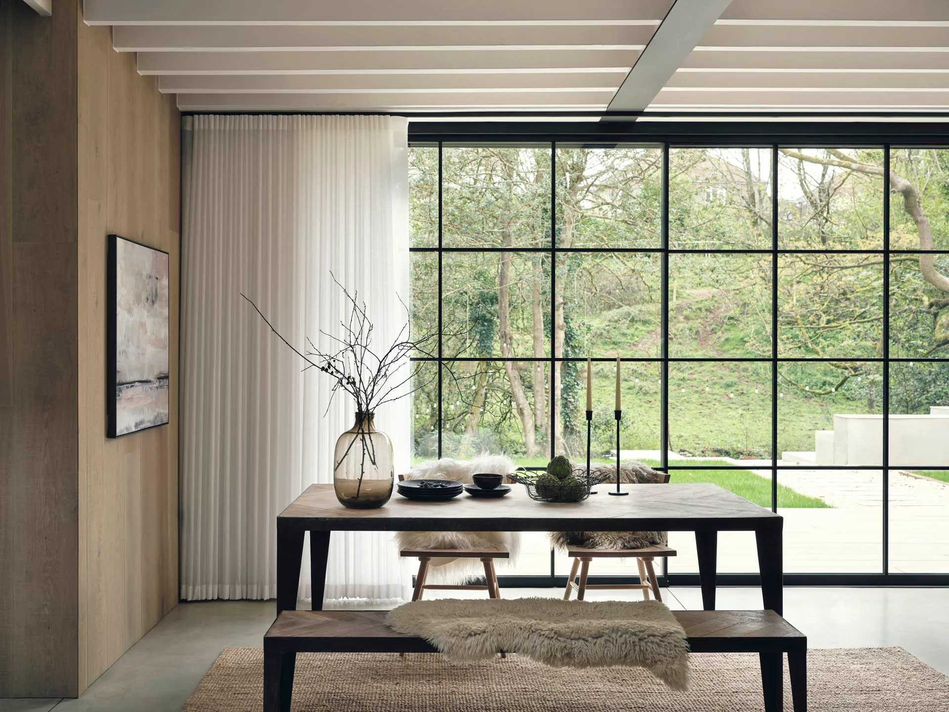 Dining table and bench beside floor-to-ceiling black-framed glazing, with white sheer wave curtains drawn back to a wooded garden.