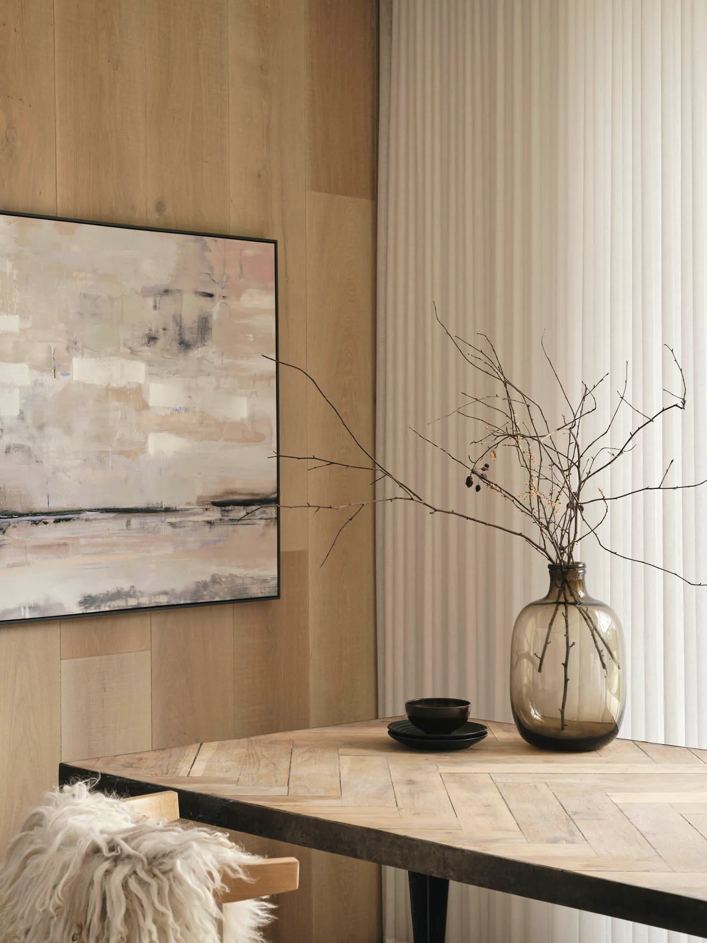 Corner of a dining room with white sheer wave curtains, a timber-panelled wall, abstract artwork and a glass vase of branches on the table.
