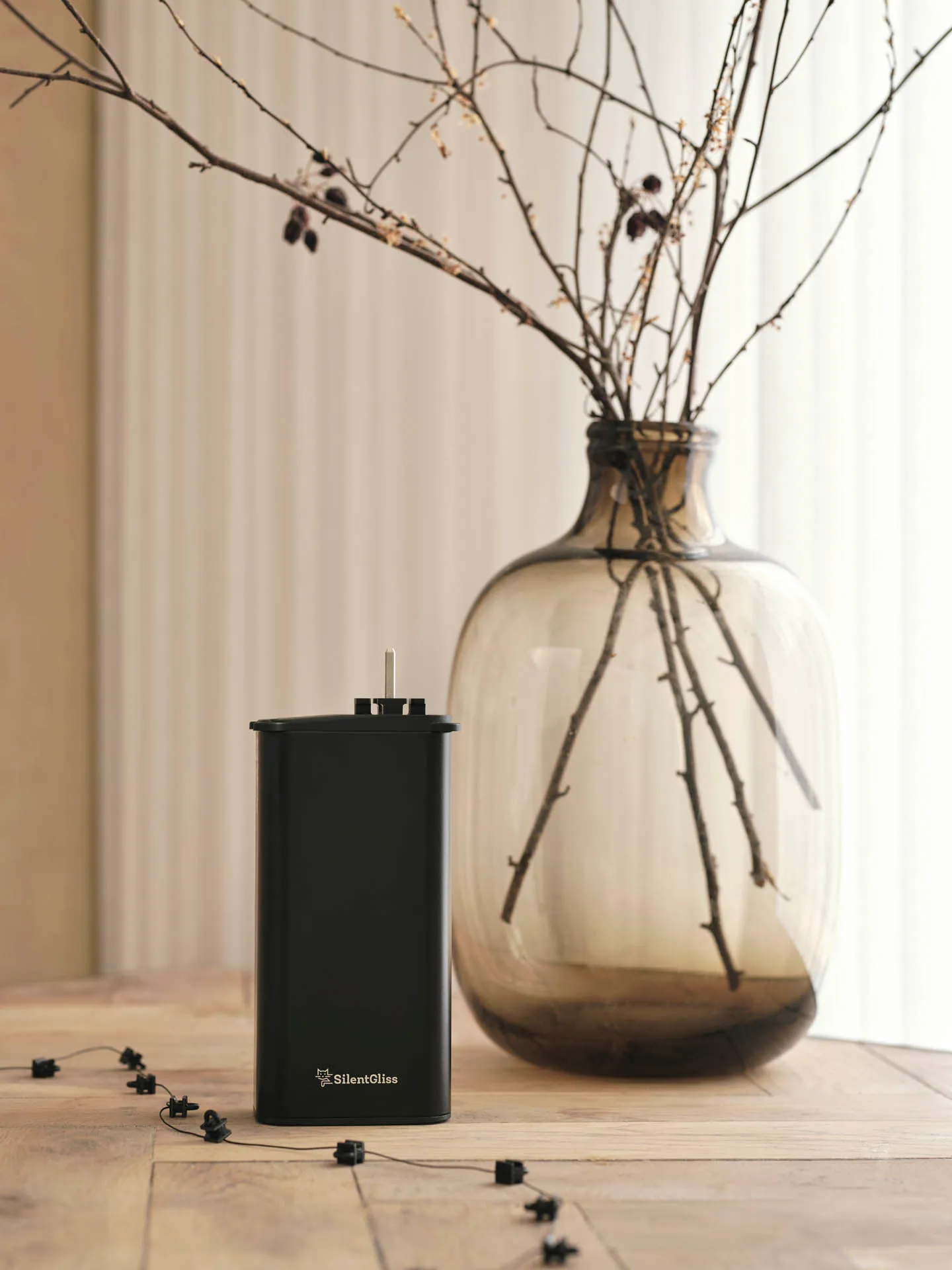 Black Silent Gliss motor unit and wave gliders on a timber table, beside a smoked-glass vase of dried branches.